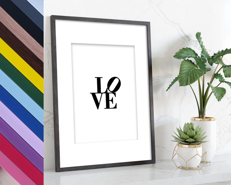May include: A black framed print with the word "LOVE" in bold, black letters. The print is hanging on a white marble wall with a green plant in a white pot and a succulent in a gold wire planter.