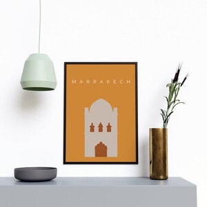 May include: A framed print with a minimalist design of a building in a muted orange color with the word "MARRAKECH" in white text at the top. The print is hanging on a white wall above a gray table with a gray bowl and a gold vase with dried grass.