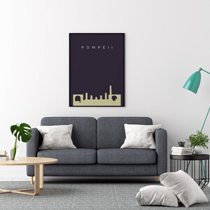 May include: A framed art print featuring the word "POMPEII" above a gold-colored skyline silhouette against a dark blue background. The print is displayed above a gray sofa with white pillows.
