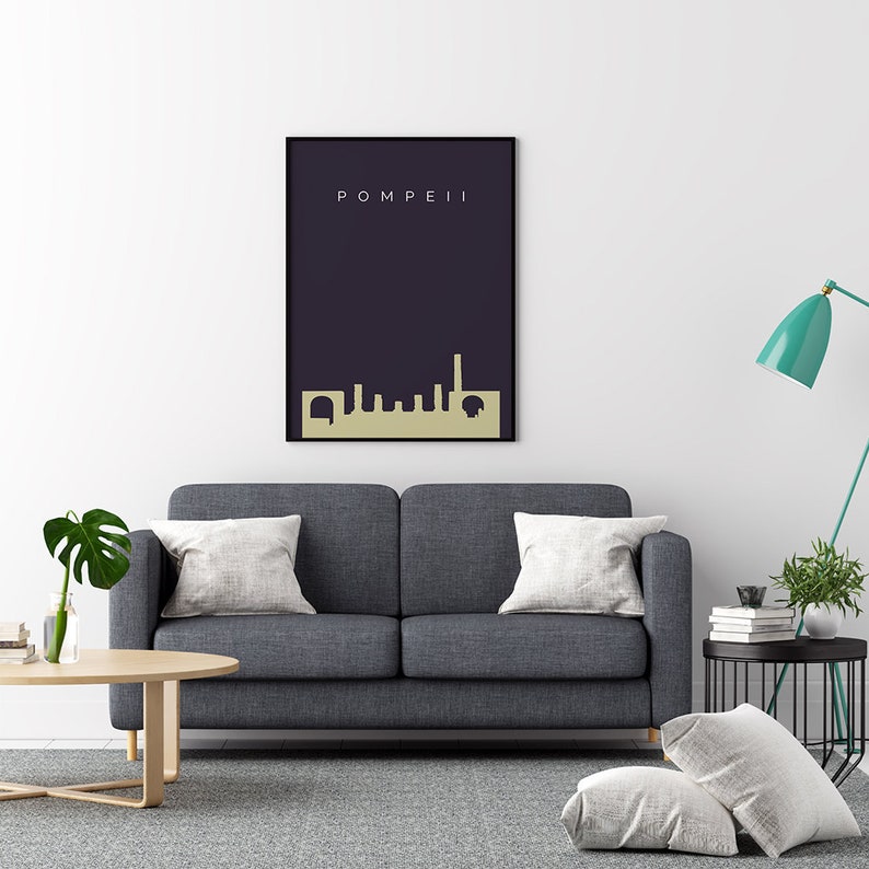 May include: A framed art print featuring the word "POMPEII" above a gold-colored skyline silhouette against a dark blue background. The print is displayed above a gray sofa with white pillows.