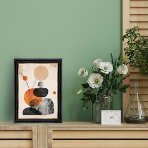May include: A black framed print with an abstract design of stacked stones and circles in orange, brown, and black against a beige background.