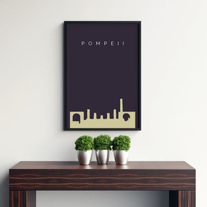 May include: A framed art print featuring the word "POMPEII" in white text above a gold-colored skyline silhouette against a dark purple background. The print is displayed on a white wall above a dark brown wooden table with three small potted plants.