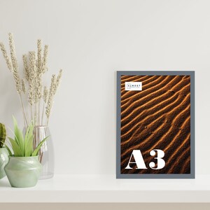 May include: A framed photograph of a desert landscape with sand dunes in shades of brown and orange. The frame is gray and has the text "Almost Always" and "A3" printed on it.