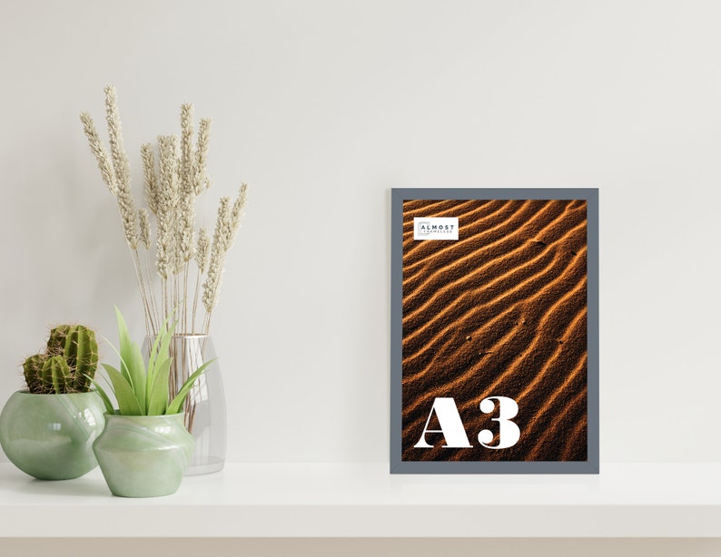 May include: A framed photograph of a desert landscape with sand dunes in shades of brown and orange. The frame is gray and has the text "Almost Always" and "A3" printed on it.