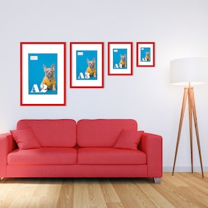 May include: Four framed prints with red borders featuring a French Bulldog in a yellow sweater on a blue backdrop. The prints are labelled A1, A2, A3, and A4. A red sofa and a wooden floor lamp are in the foreground.