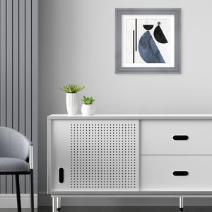 May include: A white cabinet with two drawers and a sliding door. The door has a perforated pattern. The cabinet has silver legs. A framed abstract art print with blue and black geometric shapes hangs on the wall above the cabinet.