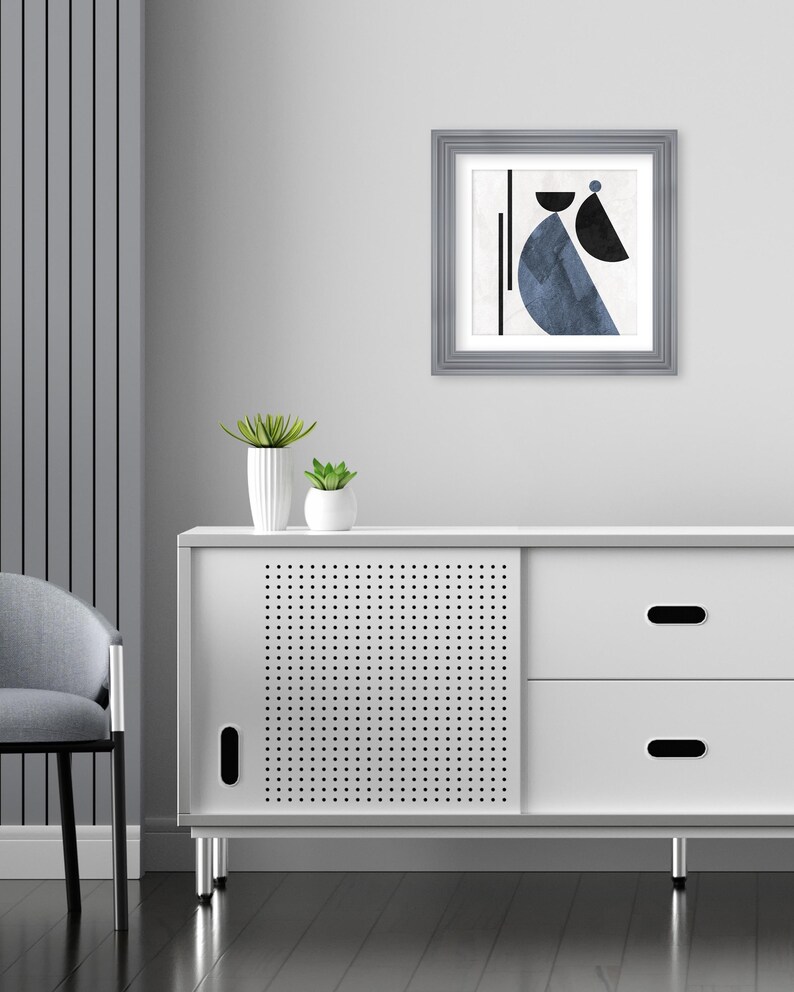 May include: A white cabinet with two drawers and a sliding door. The door has a perforated pattern. The cabinet has silver legs. A framed abstract art print with blue and black geometric shapes hangs on the wall above the cabinet.