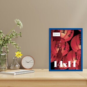 May include: A framed print with a blue border, displaying a photograph of red Chinese lanterns. The print is labeled "14x11" in white text. The frame is on a wooden surface, with a vase of flowers, a clock, and a notepad.
