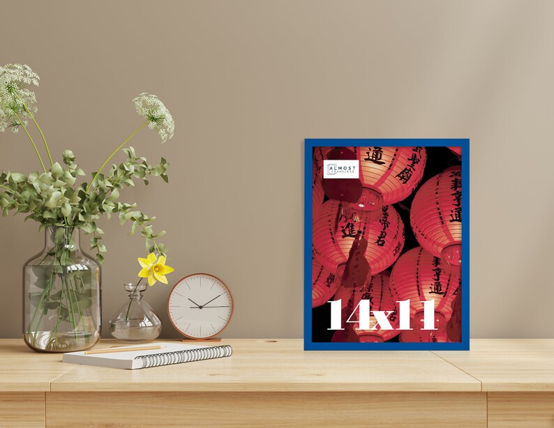 May include: A framed print with a blue border, displaying a photograph of red Chinese lanterns. The print is labeled "14x11" in white text. The frame is on a wooden surface, with a vase of flowers, a clock, and a notepad.