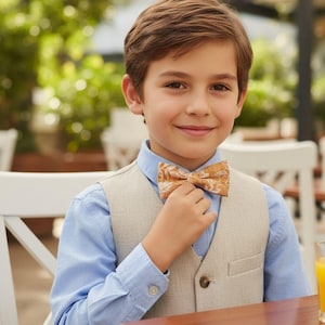 Puede incluir: Un joven con una camisa azul claro, un chaleco beige y una pajarita estampada. La pajarita presenta un diseño floral en tonos dorados y marrones. La persona sonríe, lo que sugiere una ocasión formal o de celebración.