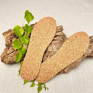 Può includere: Due solette in sughero con superficie strutturata, esposte su un pezzo di corteccia con foglie verdi. Le solette sono marroni chiare e progettate per essere inserite nelle scarpe.
