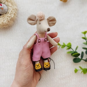 May include: A small beige fabric mouse doll featuring a light pink corduroy overall with thin straps and metal buckles. The mouse has a small beige pom-pom on its head and wears yellow shoes with black soles and a black smiley face graphic on each.