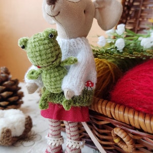May include: A plush toy mouse wearing a white knitted sweater and a red skirt, holding a green frog plush toy. The mouse is wearing red shoes with white polka dots. The toys are sitting in front of a wicker basket with yarn.