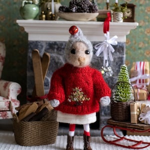 May include: A handmade doll dressed for the holidays. The doll wears a red sweater with a gold embroidered Christmas tree, a white skirt, and striped stockings. It has a gray hat with a red bell and brown boots. The doll is standing in front of a fireplace.