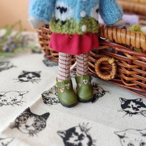 May include: A doll wearing a blue, green, and white sweater, a red skirt, and green shoes with silver hearts. The doll is sitting in a wicker basket.