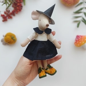Può includere: Una bambola di topo in feltro fatta a mano che indossa un cappello da strega nero, un vestito bianco con un colletto nero e scarpe di zucca gialle. La bambola è tenuta in una mano su uno sfondo bianco.