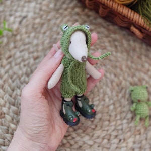 Op de afbeelding: Een klein groen kikker knuffeldier met een groene gebreide hoodie en groene laarzen. Het speelgoed wordt in de hand van een persoon gehouden.