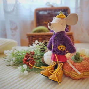 Puede incluir: Un rat&oacute;n de peluche con un su&eacute;ter de punto morado con un dise&ntilde;o de calabaza, una falda roja y botas amarillas. El rat&oacute;n sostiene una calabaza de punto.