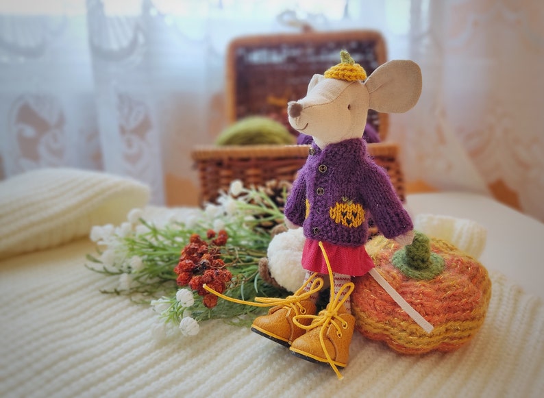 Puede incluir: Un rat&oacute;n de peluche con un su&eacute;ter de punto morado con un dise&ntilde;o de calabaza, una falda roja y botas amarillas. El rat&oacute;n sostiene una calabaza de punto.