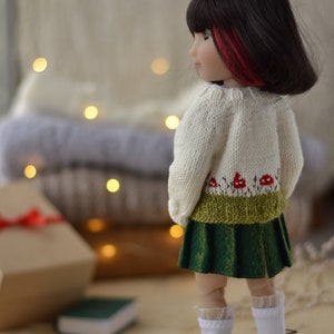 Op de afbeelding: Een pop gekleed in een cr&egrave;mekleurige gebreide trui met een paddenstoelontwerp, een groene rok, witte sokken en bruine schoenen. De pop heeft zwart haar met rode highlights. Een geschenkdoos en een klein groen boek staan op de achtergrond.