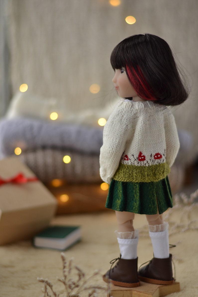 Op de afbeelding: Een pop gekleed in een cr&egrave;mekleurige gebreide trui met een paddenstoelontwerp, een groene rok, witte sokken en bruine schoenen. De pop heeft zwart haar met rode highlights. Een geschenkdoos en een klein groen boek staan op de achtergrond.