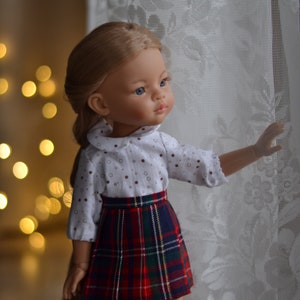 May include: A doll with long blonde hair wearing a white shirt with brown polka dots and a red and green plaid skirt. The doll is looking to the right and has blue eyes.