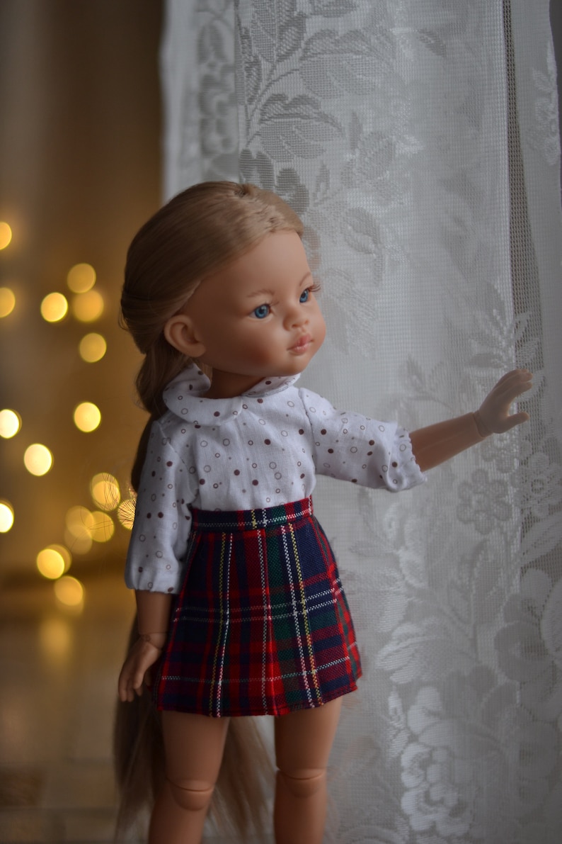 May include: A doll with long blonde hair wearing a white shirt with brown polka dots and a red and green plaid skirt. The doll is looking to the right and has blue eyes.