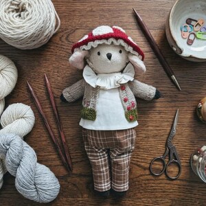 May include: A plush animal doll wearing a red and white mushroom-patterned hat, a white ruffled collar, a beige knit cardigan with small red buttons, and brown plaid trousers. The doll has a cream-colored face and is surrounded by yarn and tools.