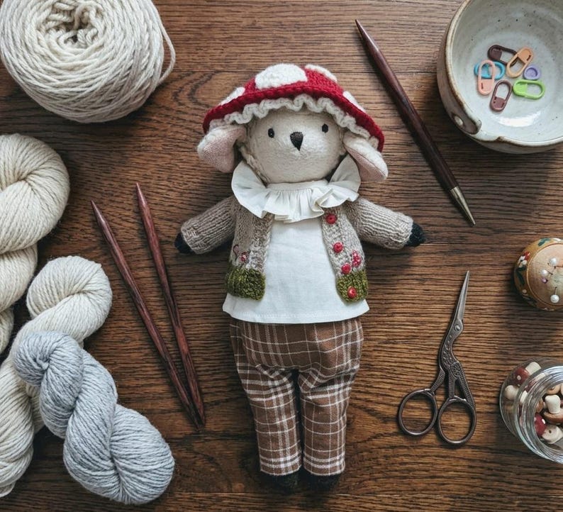 May include: A plush animal doll wearing a red and white mushroom-patterned hat, a white ruffled collar, a beige knit cardigan with small red buttons, and brown plaid trousers. The doll has a cream-colored face and is surrounded by yarn and tools.