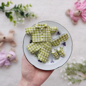 Puede incluir: Un conjunto de pijama de vichy verde y blanco para una muñeca, que incluye una camisa, pantalones y una máscara para dormir. El conjunto se muestra en un plato blanco con diseños florales.
