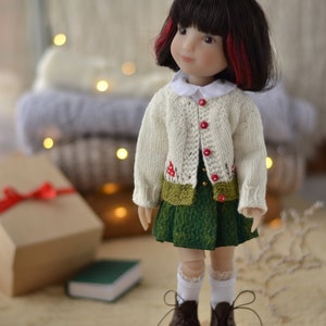 Op de afbeelding: Pop met een cr&egrave;mekleurig vest met rode knopen en een groene rok. De pop heeft zwart haar met rode highlights en bruine schoenen. De pop staat op een houten stervormige basis.