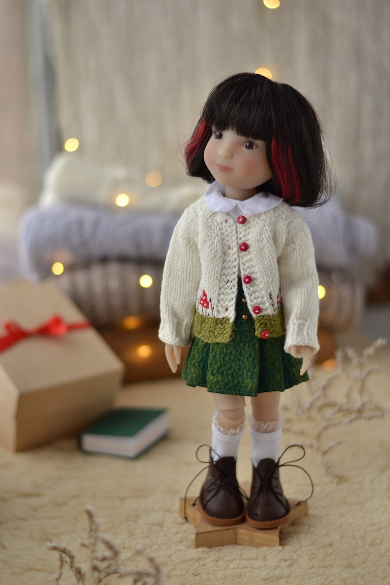 Op de afbeelding: Pop met een cr&egrave;mekleurig vest met rode knopen en een groene rok. De pop heeft zwart haar met rode highlights en bruine schoenen. De pop staat op een houten stervormige basis.