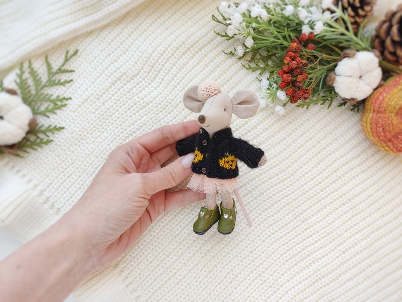 Puede incluir: Un peque&ntilde;o rat&oacute;n de peluche que lleva un c&aacute;rdigan de punto negro con detalles de calabaza amarilla y una falda de tul rosa. El rat&oacute;n tiene zapatos verdes y un lazo rosa en la cabeza.