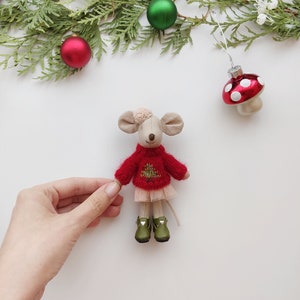 Op de afbeelding: Een kleine, witte, pluche muis die een rode trui draagt met een groen kerstboomontwerp en een roze tule rok. De muis draagt groene laarzen met witte hartjes. De muis zit op een wit oppervlak met groene dennentakken en rode en groene ornamenten.