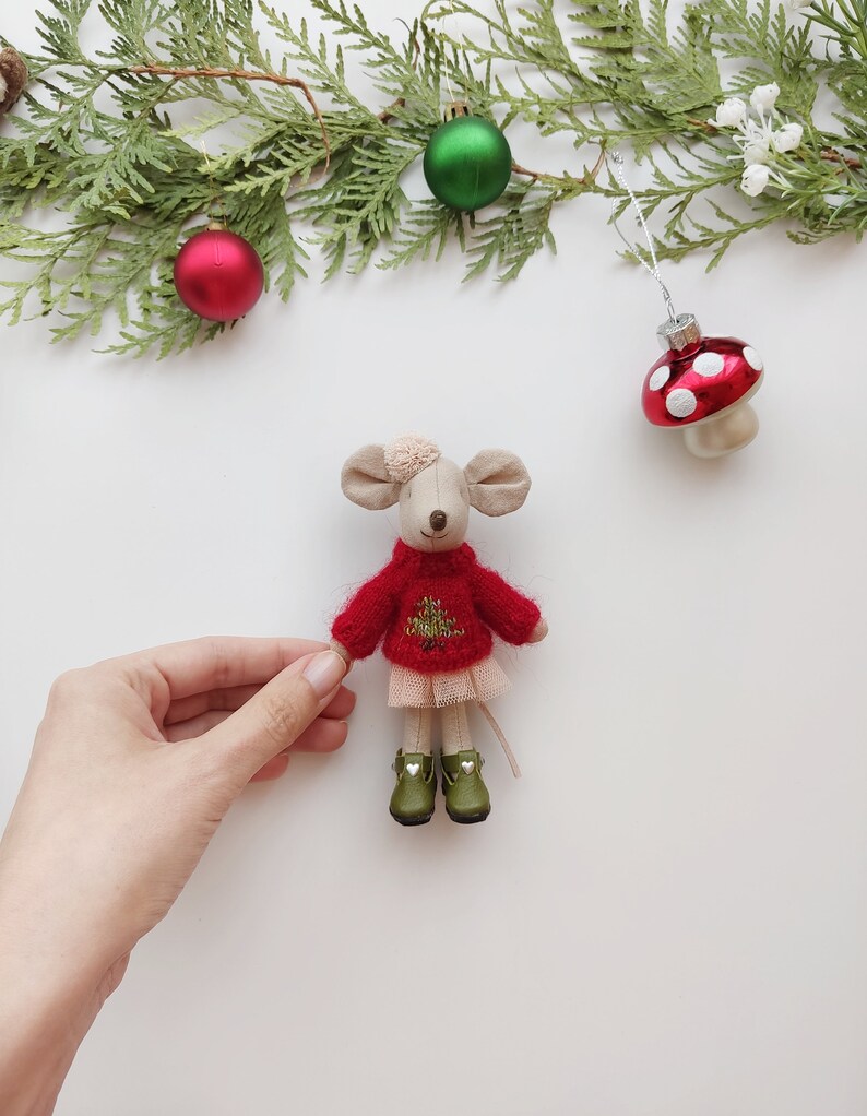 Op de afbeelding: Een kleine, witte, pluche muis die een rode trui draagt met een groen kerstboomontwerp en een roze tule rok. De muis draagt groene laarzen met witte hartjes. De muis zit op een wit oppervlak met groene dennentakken en rode en groene ornamenten.