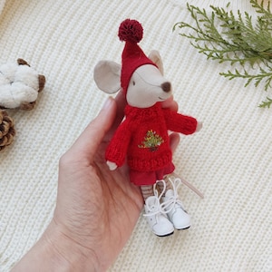 Op de afbeelding: Een klein grijs muis knuffeldier met een rode trui met een groen kerstboomontwerp en witte laarzen. De muis houdt een kleine rood-wit gestreepte sjaal vast.