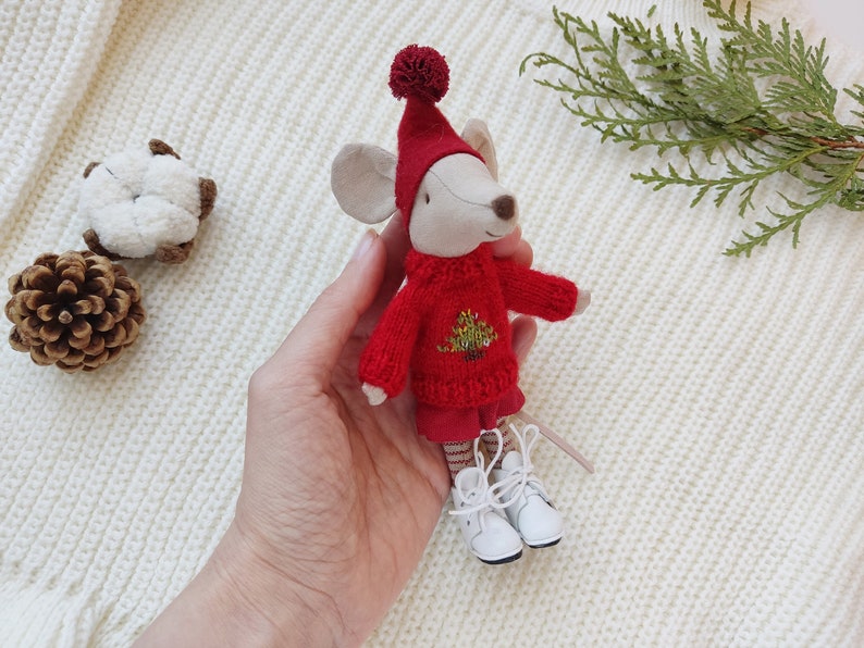 Op de afbeelding: Een klein grijs muis knuffeldier met een rode trui met een groen kerstboomontwerp en witte laarzen. De muis houdt een kleine rood-wit gestreepte sjaal vast.