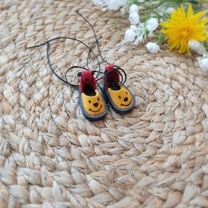 May include: A pair of yellow felt shoes with black laces and a smiling face design. The shoes are on a woven straw mat.