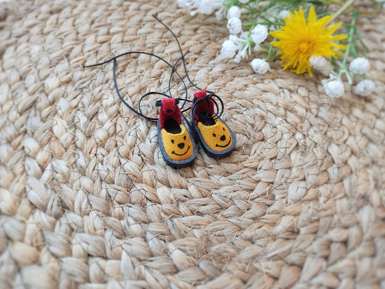 May include: A pair of yellow felt shoes with black laces and a smiling face design. The shoes are on a woven straw mat.