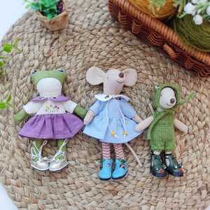 May include: Three handmade plush toys: a frog wearing a purple dress, a mouse wearing a blue dress, and a green frog wearing a green knitted outfit. The toys are sitting on a woven straw mat.