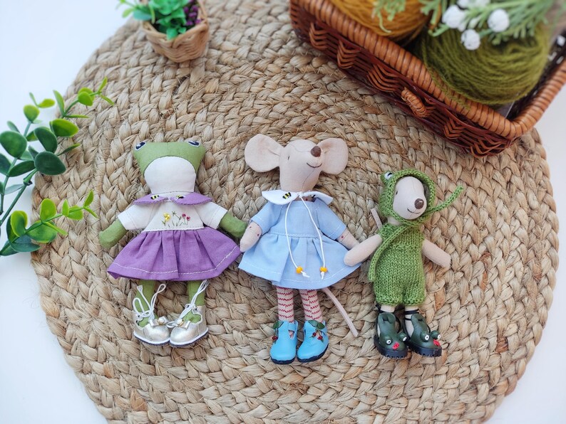May include: Three handmade plush toys: a frog wearing a purple dress, a mouse wearing a blue dress, and a green frog wearing a green knitted outfit. The toys are sitting on a woven straw mat.