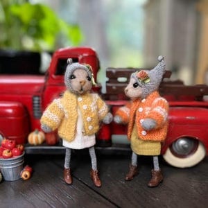 May include: Two small, gray, knitted animal figurines wearing sweaters and standing in front of a red toy truck. The figurine on the left is wearing a yellow sweater with white accents and the figurine on the right is wearing an orange sweater with white accents.