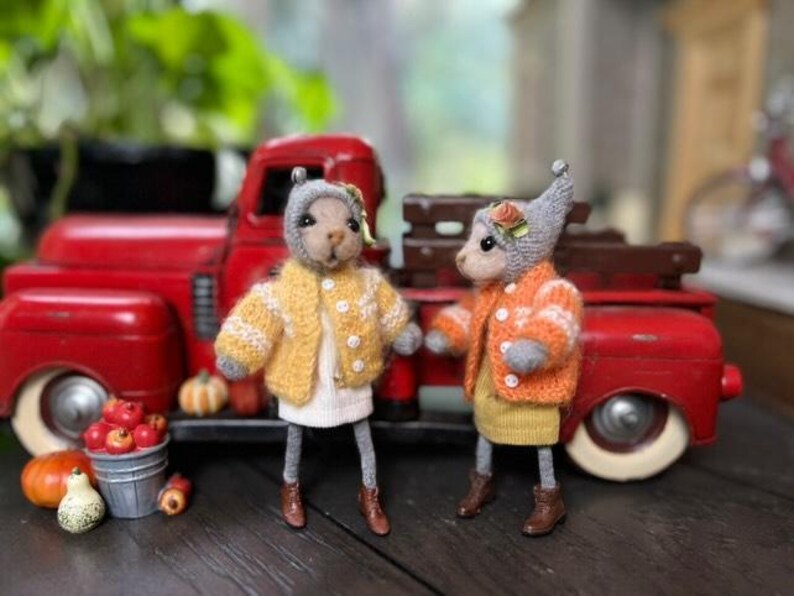 May include: Two small, gray, knitted animal figurines wearing sweaters and standing in front of a red toy truck. The figurine on the left is wearing a yellow sweater with white accents and the figurine on the right is wearing an orange sweater with white accents.