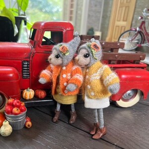 May include: Two small knitted plush toys dressed in orange and yellow sweaters with grey hats and brown boots. The toys are standing in front of a red toy truck with a wooden bed.