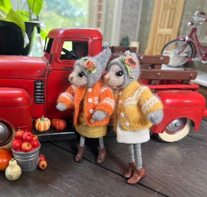 May include: Two small knitted plush toys dressed in orange and yellow sweaters with grey hats and brown boots. The toys are standing in front of a red toy truck with a wooden bed.