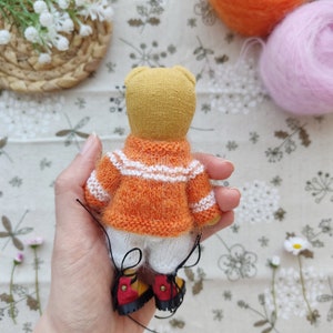 May include: A small knitted teddy bear wearing an orange and white striped sweater and white pants. The bear is wearing red and yellow shoes with black laces. The bear is being held by a hand.