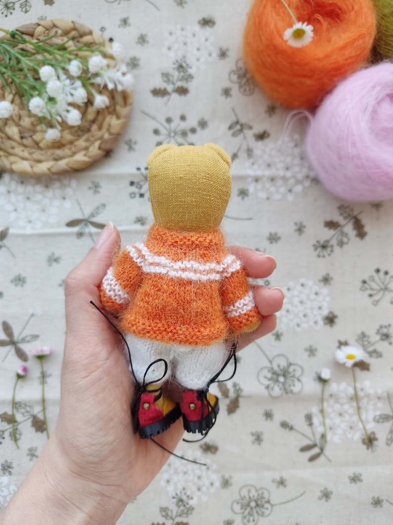 May include: A small knitted teddy bear wearing an orange and white striped sweater and white pants. The bear is wearing red and yellow shoes with black laces. The bear is being held by a hand.