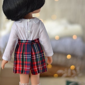 May include: A doll wearing a white collared shirt with long sleeves and a red, black, and white plaid skirt.
