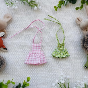 Puede incluir: Dos vestidos de muñeca en miniatura, uno rosa a cuadros y otro verde floral, con tirantes. Los vestidos están rodeados de peluches de animales, como un zorro, un ratón y un erizo, sobre una superficie de punto blanca.