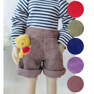 May include: A doll wearing a striped long-sleeved shirt and brown corduroy shorts. The doll is holding a yellow stuffed animal with a red heart.